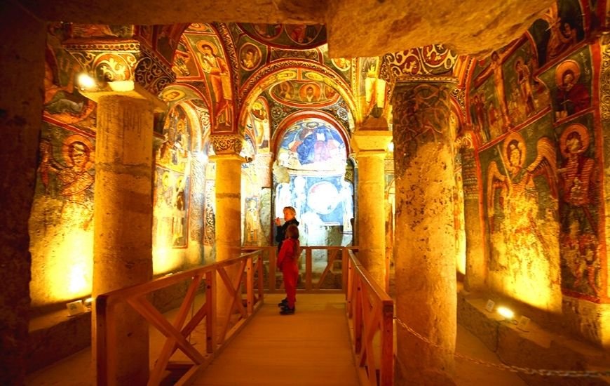 Interior iluminado de iglesia rupestre con frescos bizantinos y visitantes observando detalles históricos en Capadocia