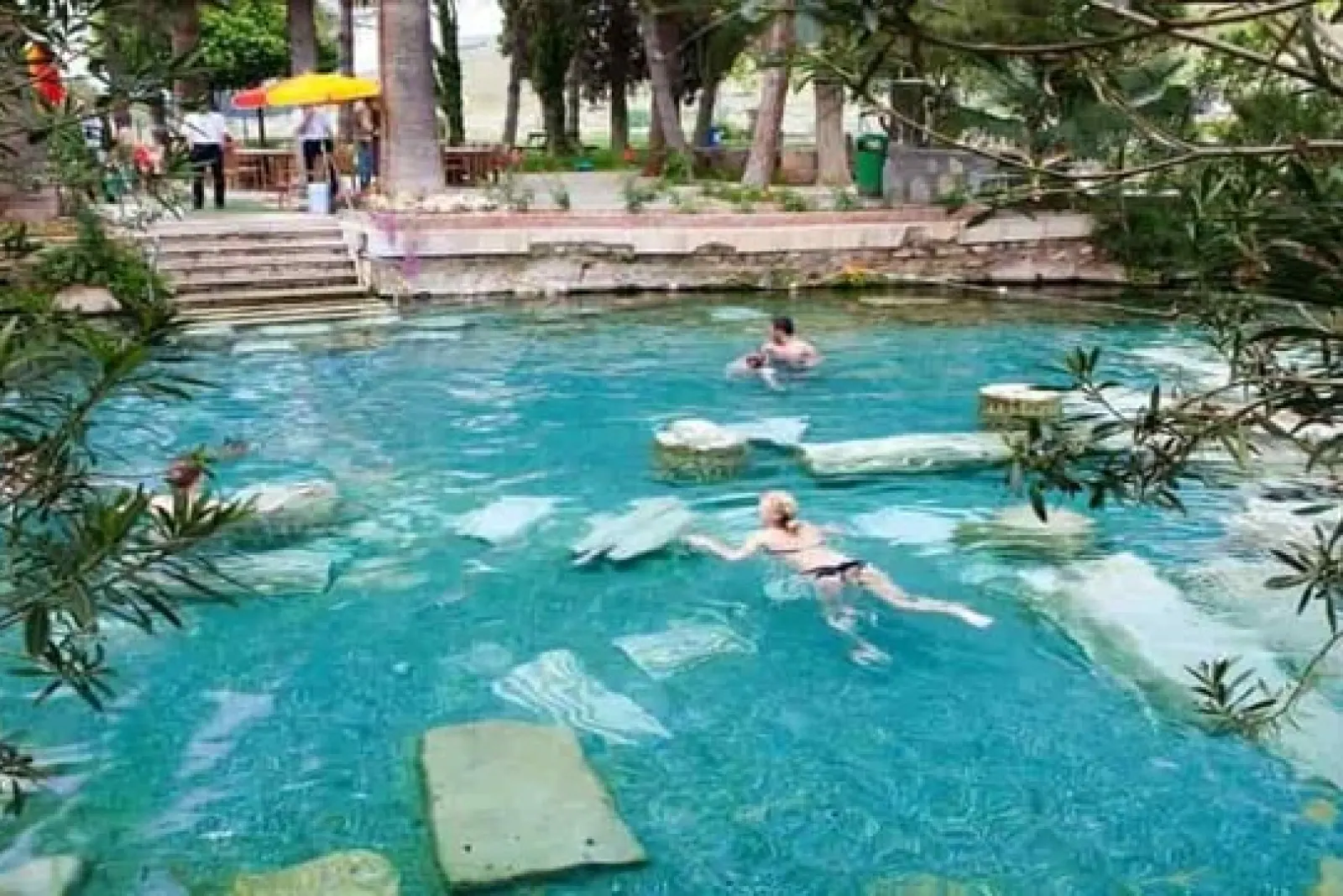 Personas nadando en una piscina termal con ruinas antiguas bajo el agua y vegetación alrededor