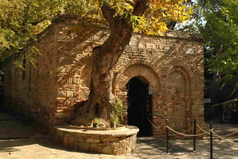 Vista exterior de la Casa de la Virgen María, un antiguo edificio de piedra con arcos en un patio soleado y arbolado.