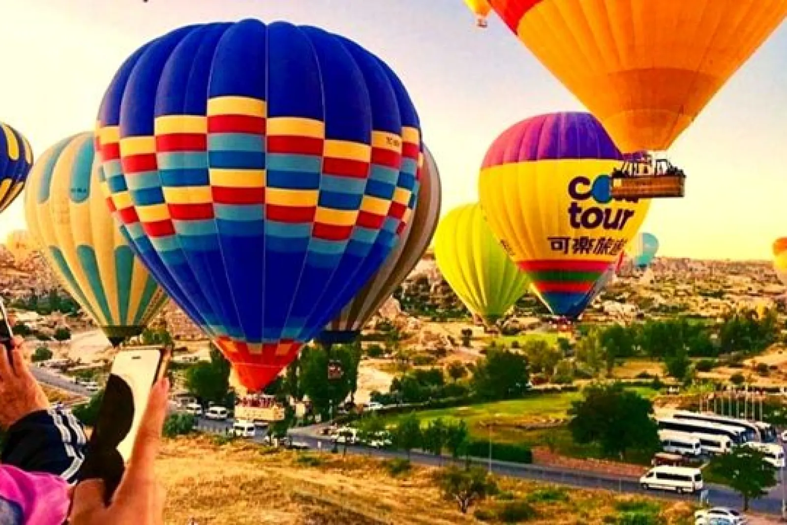 Globos aerostáticos de colores ascendiendo sobre el paisaje de Capadocia al amanecer, con terreno semiárido y árboles dispersos.