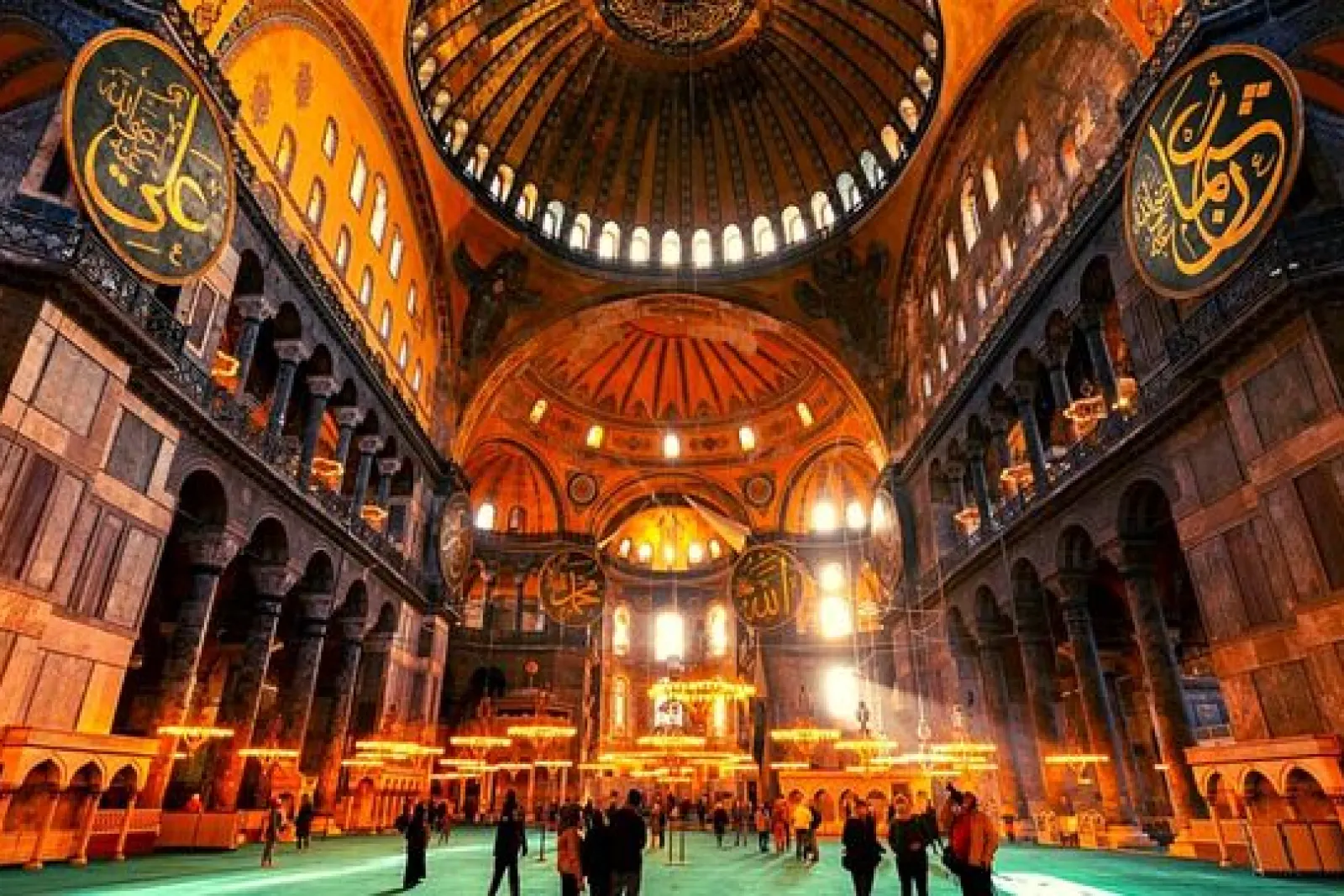 Interior de Santa Sofía en Estambul con grandes cúpulas iluminadas y visitantes sobre la alfombra verde.